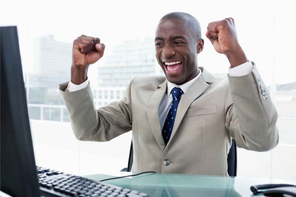 man sitting in front of computer smiling and raising arms in celebration