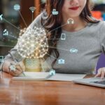 A woman writing in a notebook while sitting at a desk, with a laptop nearby. Holographic icons of a brain, devices, and technology-related symbols float above her notebook, representing AI and its applications in various fields