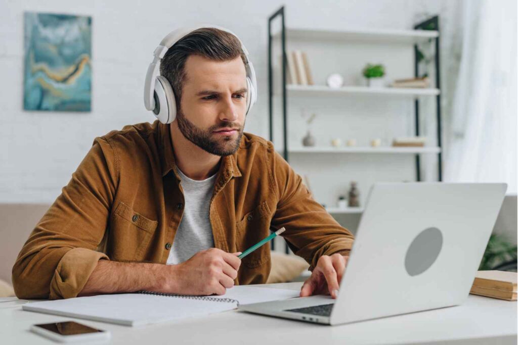 A man with headphones is sitting at a desk, focused on a laptop screen while holding a pencil. Notebooks, a smartphone, and other study materials are spread out on the desk in a home office setting