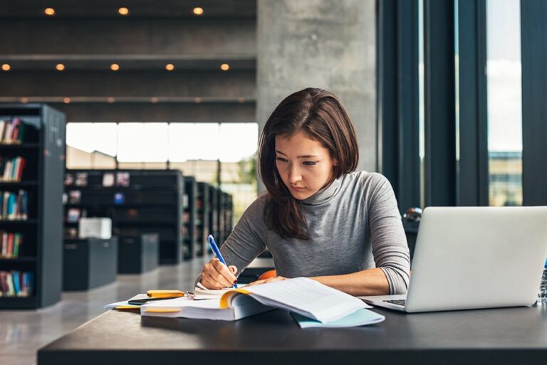 Woman studying practice exams for certification success with books and laptop in a library setting.