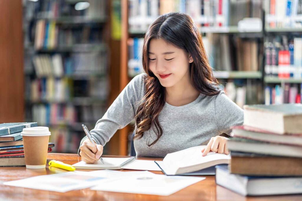 A young woman with long hair is sitting at a wooden table in a library, writing in a notebook while reading a book. She appears focused, with a stack of books next to her and a coffee cup on the table