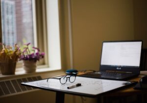 Study setup with a laptop, glasses, whiteboard, and marker on a desk next to a window.